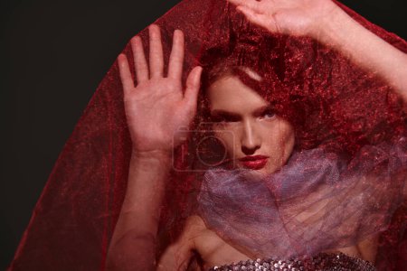 Eine junge Frau mit auffallend roten Haaren posiert elegant in einem Studio-Setting und trägt einen Schleier auf dem Kopf.