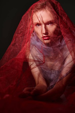 A young woman exudes classic beauty in a stunning red veil and dress, striking a pose in a studio setting against a black backdrop.
