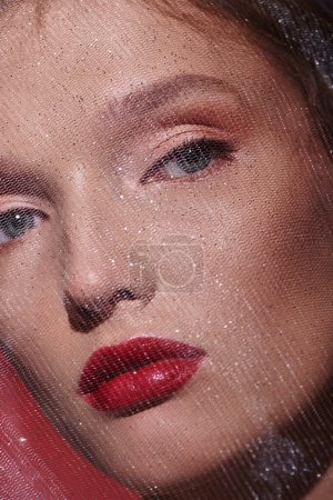 Una joven con una belleza clásica, con lápiz labial rojo, mira seductoramente en una foto de cerca sobre un fondo negro.
