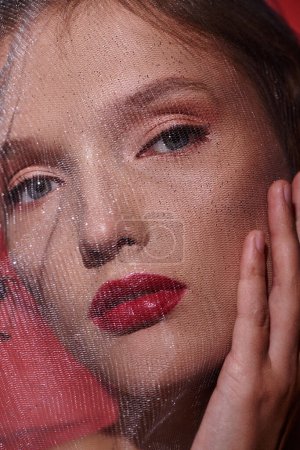 Eine junge Frau strahlt klassische Schönheit mit rotem Lippenstift und einem Schleier aus, der ihr Gesicht verhüllt, als sie in einem Studio posiert.
