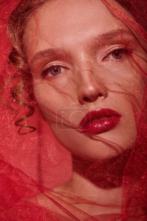 Une jeune femme respire la beauté classique alors qu'elle pose avec un voile rouge ornant sa tête dans un décor de studio.
