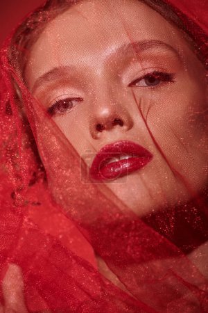 Une jeune femme respire l'élégance dans un décor studio, ornée d'un voile rouge saisissant et d'un rouge à lèvres rouge audacieux.