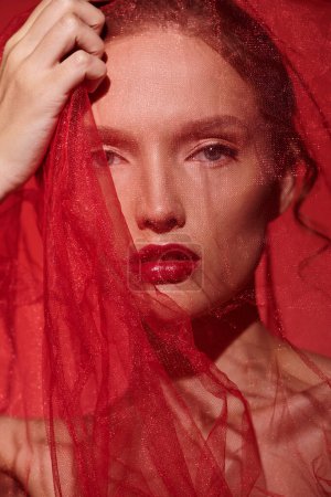 Une jeune femme respire la beauté classique, ses cheveux roux en cascade sous un voile rouge saisissant dans un décor de studio sur fond noir.