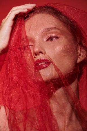 Une jeune femme respire la beauté classique, la tête ornée d'un voile rouge saisissant dans un décor studio sur fond noir.