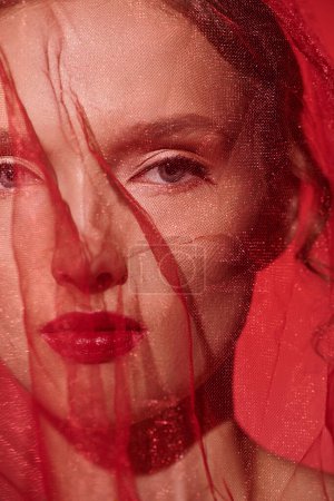 Une jeune femme aux cheveux roux saisissants pose dans un studio, le visage partiellement obscurci par un voile délicat.