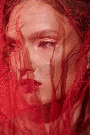 A young woman with striking red hair poses elegantly with a veil covering her face, exuding classic beauty in a studio setting.
