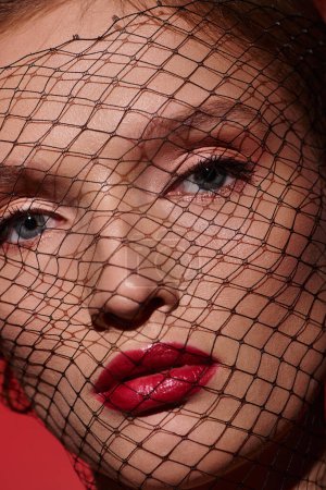 A young woman exudes classic beauty with striking red lipstick, her face partially covered by an intricate net veil.