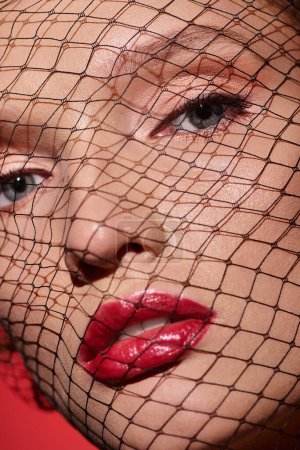 Una joven con una belleza clásica usa lápiz labial rojo y una red que cubre su rostro en una pose cautivadora y misteriosa..