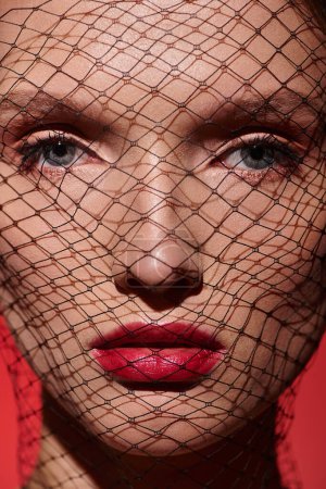Une jeune femme à la beauté classique pose dans un studio avec un voile rouge couvrant son visage, respirant mystère et élégance.
