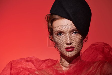 Eine junge Frau strahlt klassische Schönheit in einem auffallend roten Kleid und schwarzem Hut aus, als sie selbstbewusst in einem Studio-Setting posiert.