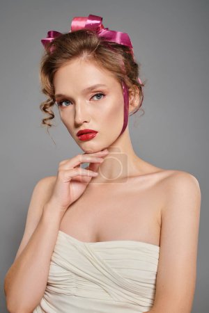 A young woman exudes classic beauty in a white dress and a pink bow on her head while posing gracefully in a studio setting.