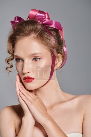 Una joven con un lazo rosado en el pelo posa graciosamente en un estudio, exudando belleza clásica sobre un fondo gris.