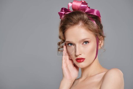 A young woman exudes classic beauty while posing in a studio, wearing a pink bow on her head against a grey backdrop.