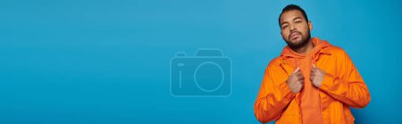 horizontale Aufnahme eines attraktiven afrikanisch-amerikanischen jungen Mannes in orangefarbenem Outfit auf blauem Hintergrund