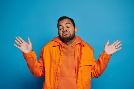 Photo for Charismatic african american man in orange outfit showing that knows nothing on blue background - Royalty Free Image