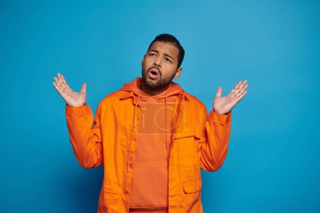 verwirrter afrikanisch-amerikanischer Mann in orangefarbenem Outfit, der auf blauem Hintergrund die Hände zur Seite legt