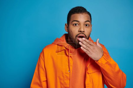 surprised african american man in orange outfit covering mouth with hand on blue background