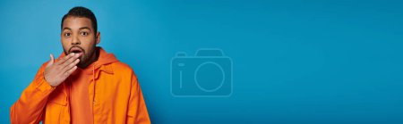 banner of amazed african american man in orange outfit covering mouth with hand on blue background
