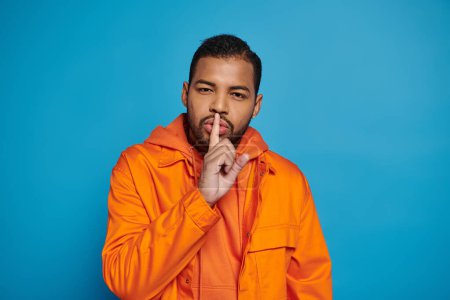 attractive african american man in orange outfit showing hush gesture silence on blue background