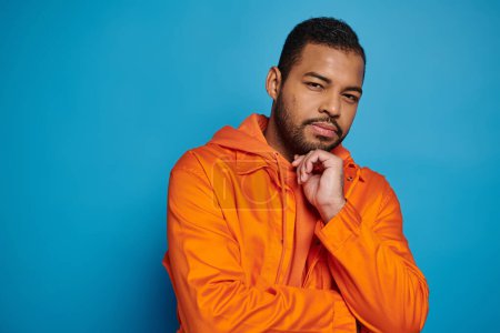 charismatic african american man in orange outfit touching to chin with hand on blue background