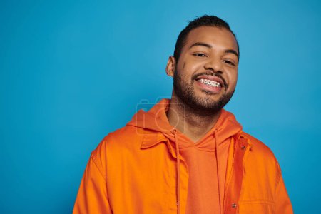 Téléchargez les photos : Homme afro-américain joyeux dans la vingtaine souriant largement avec la tête penchée sur fond bleu - en image libre de droit