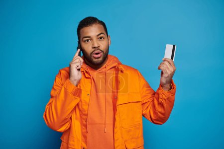 surprised african american young guy calling in smartphone with credit card on blue background