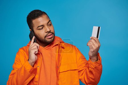 Foto de Charming african american man in orange outfit calling in smartphone and looking to credit card - Imagen libre de derechos