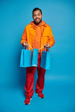 Téléchargez les photos : Cheerful african american man in orange outfit holding shopping bags against blue background - en image libre de droit