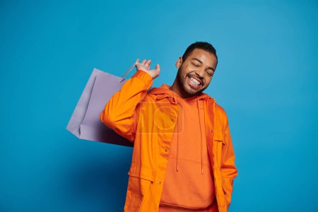 Téléchargez les photos : Joyeux homme afro-américain en tenue orange posant avec sac à provisions sur l'épaule et les yeux fermés - en image libre de droit