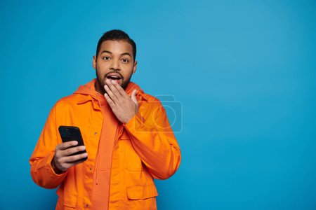 Téléchargez les photos : Surpris homme afro-américain en tenue orange défilant dans les médias sociaux et couvrant la main à la bouche - en image libre de droit