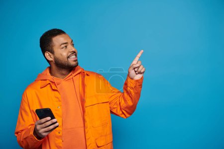 Charmanter afrikanisch-amerikanischer junger Mann in orangefarbenem Outfit mit Smartphone, das den Finger nach oben zeigt