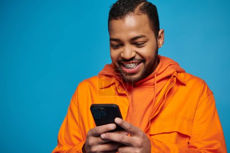 alegre afroamericano hombre en traje naranja jugando juego en teléfono inteligente sobre fondo azul