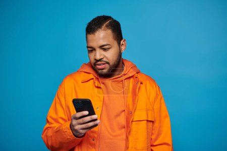 confused african american young guy in orange outfit scrolling in social media on blue background