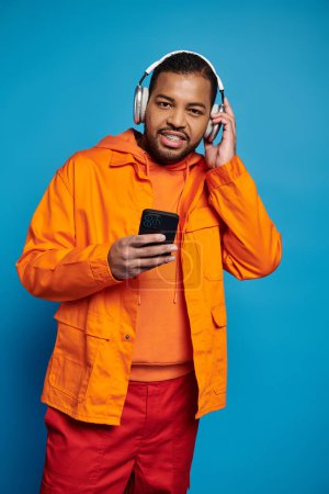 Téléchargez les photos : Charmant homme afro-américain dans les écouteurs écoutant de la musique avec smartphone et regardant à la caméra - en image libre de droit