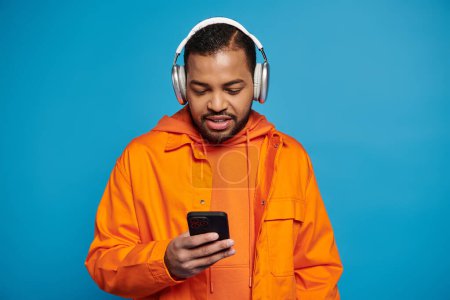 Afrikanisch-amerikanischer Typ in orangefarbenem Outfit und Kopfhörer scrollen in den sozialen Medien auf blauem Hintergrund