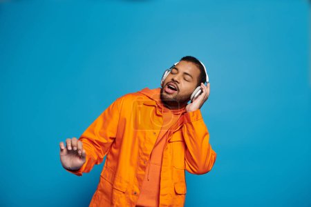 african american man in orange outfit and headphones singing with closed eyes on blue background