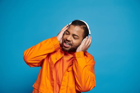 Photo for African american young man in orange outfit listening to music and holding with hands on headphones - Royalty Free Image