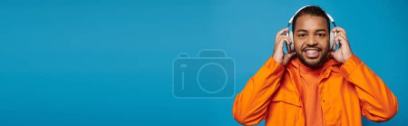 Foto de Pancarta de hombre afroamericano en traje naranja mirar a la cámara y sostener con las manos en los auriculares - Imagen libre de derechos