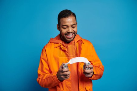 smiling african american man in orange outfit holding with hands headphones on blue background