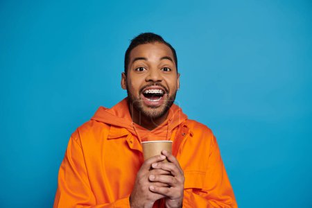 Téléchargez les photos : Cheerful african american young man in orange outfit holding with hands paper cup on blue background - en image libre de droit
