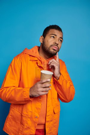 thoughtful african american man in his 20s with paper cup in hand on blue background