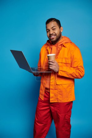 attrayant homme afro-américain en tenue orange avec tasse en papier et ordinateur portable sur fond bleu