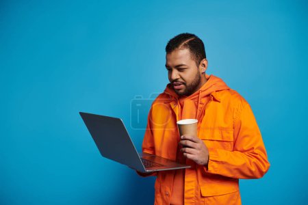 Téléchargez les photos : Beau jeune homme afro-américain en tenue orange tenant tasse en papier et ordinateur portable sur fond bleu - en image libre de droit