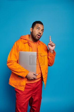 thoughtful african american man in orange outfit posing with laptop and putting finger to up