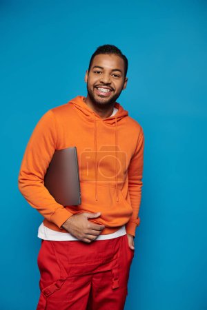 attractive african american guy in vibrant outfit posing with laptop under arm on blue background