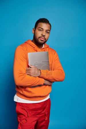 Téléchargez les photos : Charming african american man in vibrant outfit holding arms with laptop on blue background - en image libre de droit