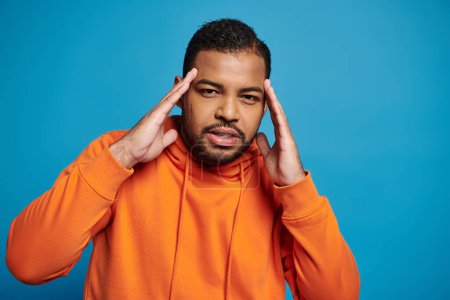thoughtful african american man in orange outfit touching to temple with hands on blue background