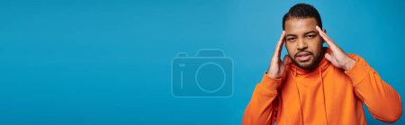 Téléchargez les photos : Bannière de l'homme afro-américain en tenue orange touchant au temple avec les mains sur fond bleu - en image libre de droit