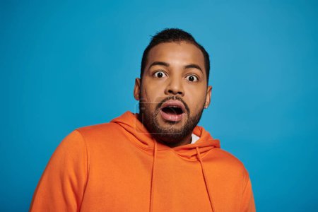 Photo for Attractive african american man in orange outfit was surprised against blue background - Royalty Free Image