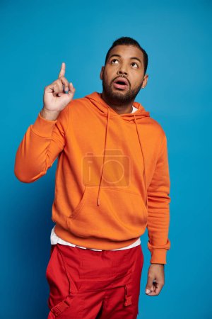 nachdenklicher afrikanisch-amerikanischer Mann in orangefarbenem Outfit, der vor blauem Hintergrund den Finger nach oben reckt